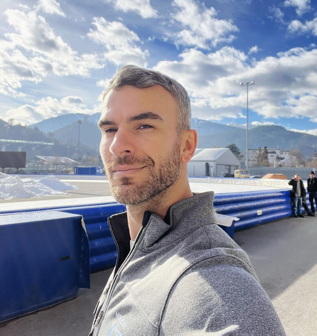 Eric Radford taking a selfie in front of an outdoor ice skating rink, and wearing a grey pullover. 