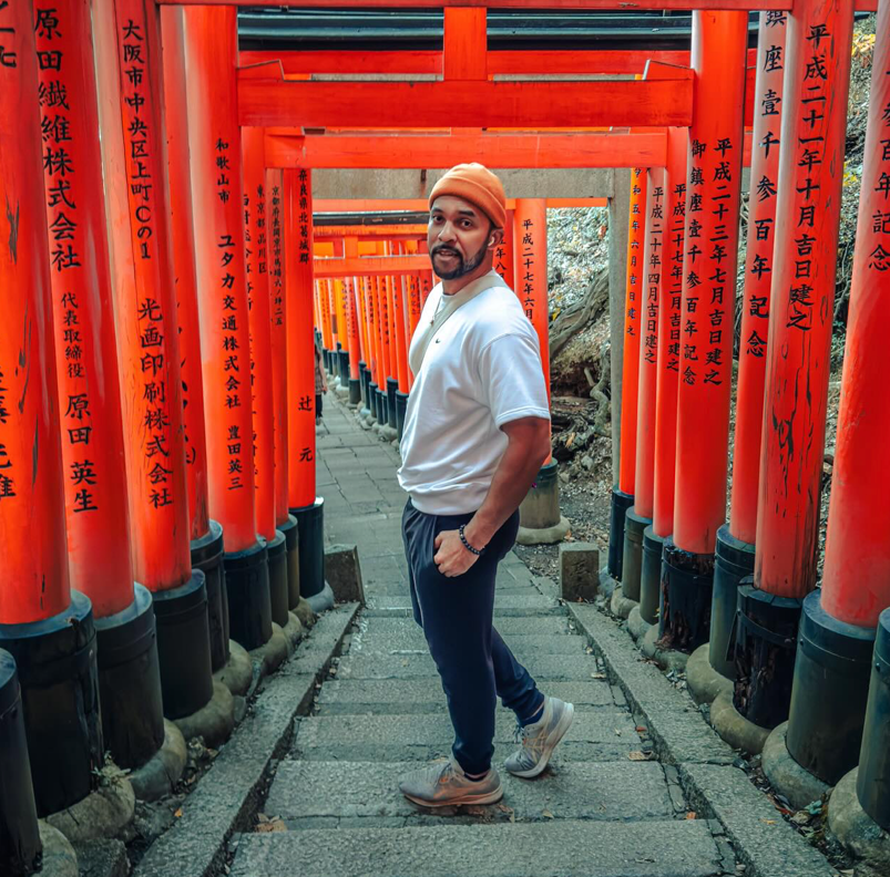Olympian Chris Kinney standing under orange poles with black Japanese lettering, wearing an orange beanie and white Lacoste shirt-sleeve shirt with AirPods in his ears.