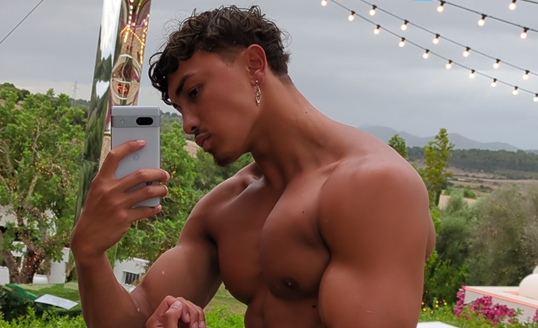 Ben Richardson, a 22-year-old Filipino man with big muscles, stands shirtless in front of a mirror taking a selfie on his gray photo while flexing. He stands outside surrounded by greenery and a pole with string lights hanging from it. He wears a diamond shaped gold earring.
