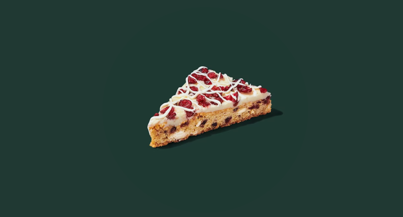 A triangle-shaped light-colored brownie bar, filled with cranberries, sits against a navy green backdrop. The dessert is covered in white cream-cheese frosting, red dried cranberries, and zig-zag icing.