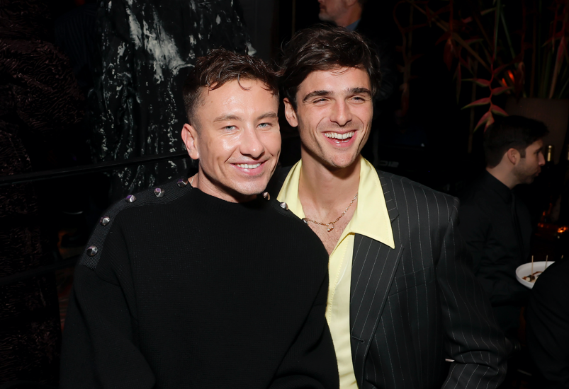 Barry Keoghan, with messy brown hair, smiles wearing a long black sweater with a mesh strip and metallic buttons affixed over his shoulders. Behind him, Jacob Elordi holds his arm around him while laughing. Elordi wears a chain around his neck and an unbuttoned pastel yellow dress shirt showing off a bit of chest hair underneath a black suit jacket with silver lines alongside it. The two stand in front of a crowd in a dark room at GQ's Men of the Year party.