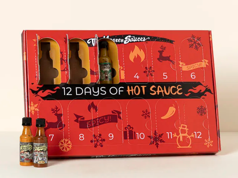 A red cardboard rectangular box is pictured from an angle in front of an off-white background. It reads "12 Days of Hot Sauce" with twelve marked doors that open up to reveal mini hot sauce bottles. Three doors are open, showing small little bottles of hot sauce with labels that are not legible.
