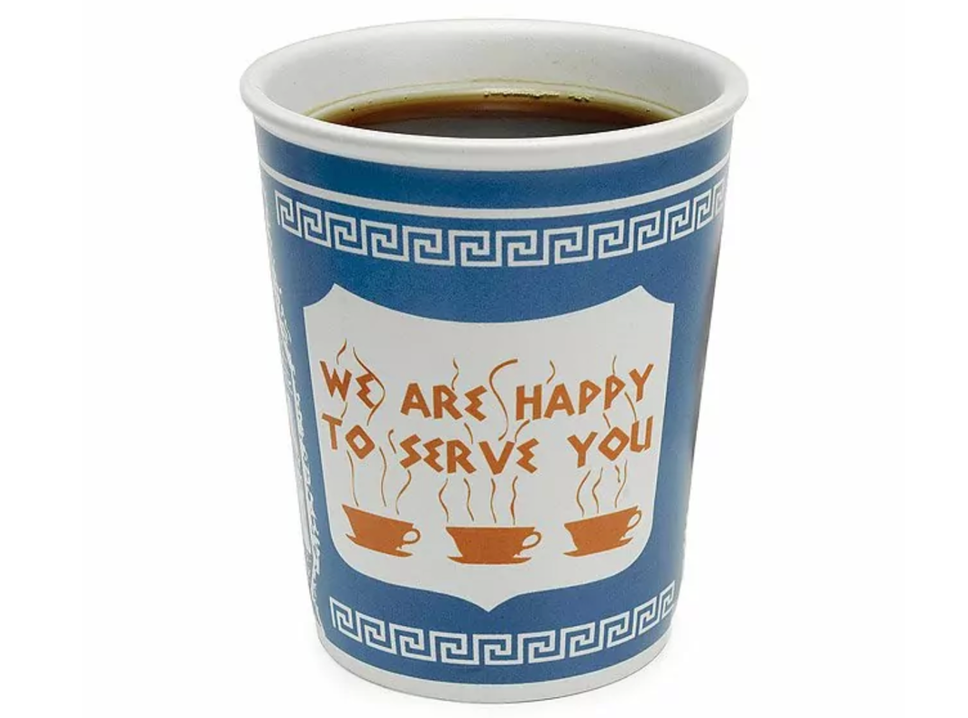 A ceramic Greek coffee cup is pictured at an angle, filled with dark coffee. The white cup has a blue border and reads "We are happy to serve you" in golden letters above illustrations of three steaming golden coffee cups.