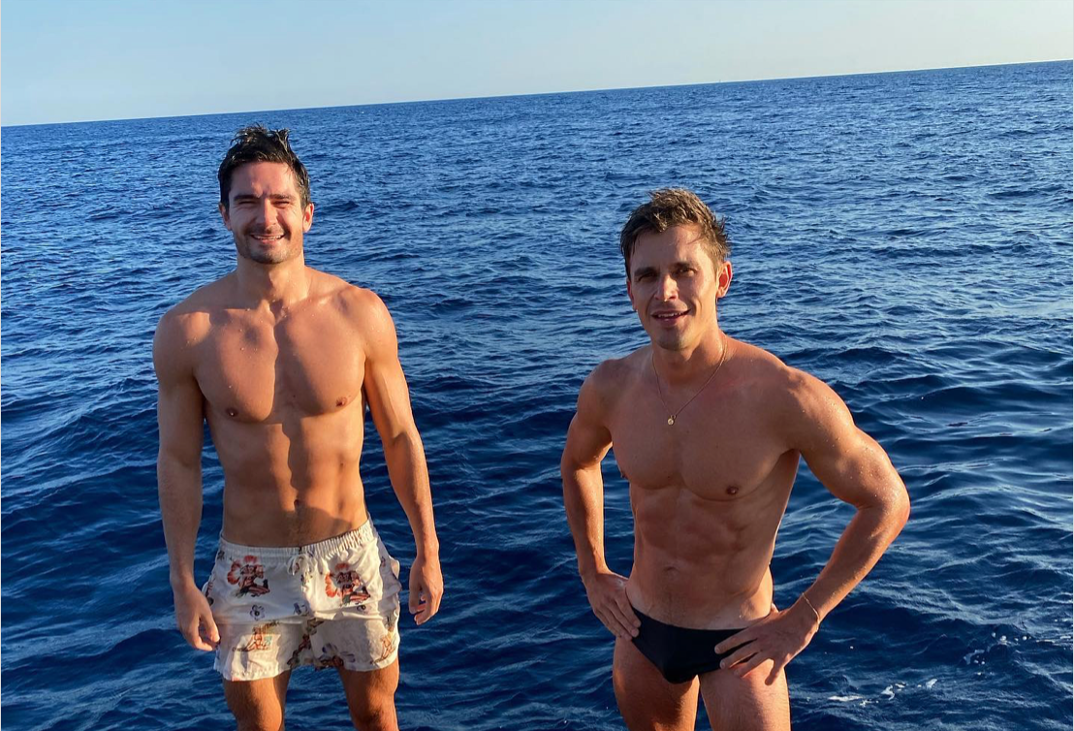 Kevin Harrington and Antoni Porowski standing on a dock in their swimsuits. 