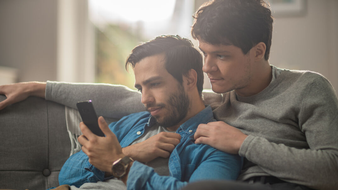 A male gay couple –– one with dark hair, a blue denim shirt, and a dark beard, and the other with dark blonde hair and a gray sweatshirt –– sit on a gray couch looking at a phone screen while in an embrace. They sit in a light house out of focus with a modern interior.