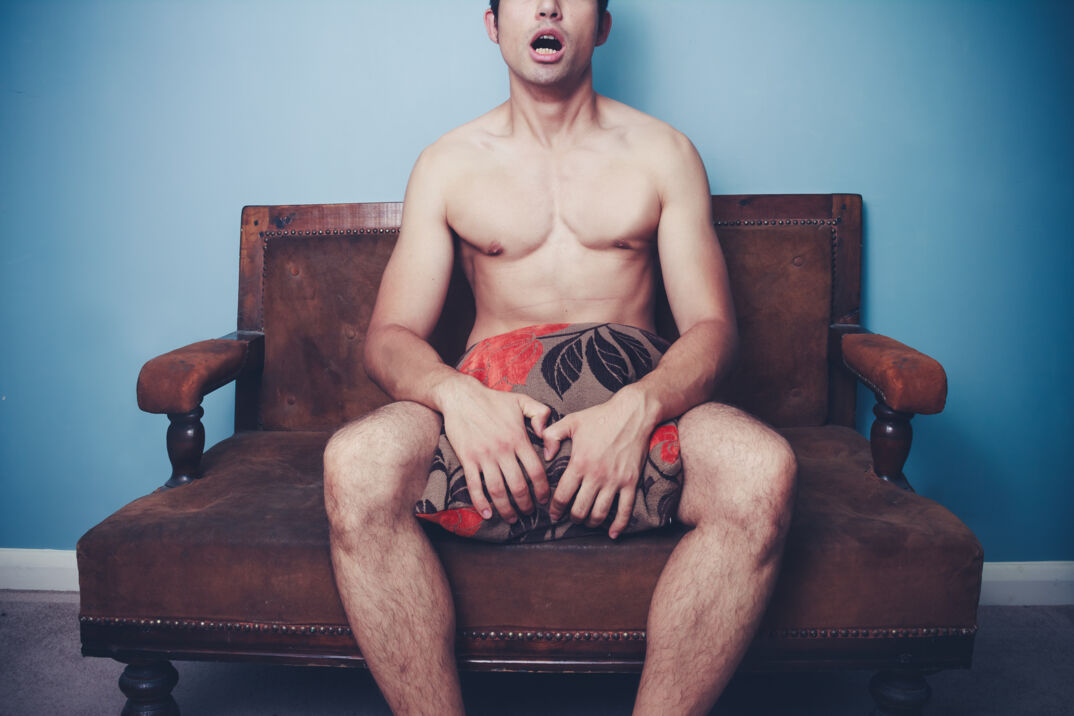 A naked young woman with hairy legs and a smooth chest sits on a brown couch covering his lower section with a decorate pillow. His mouth is open in shock and surprise and his eyes are out of frame. He sits in front of a blue wall.
