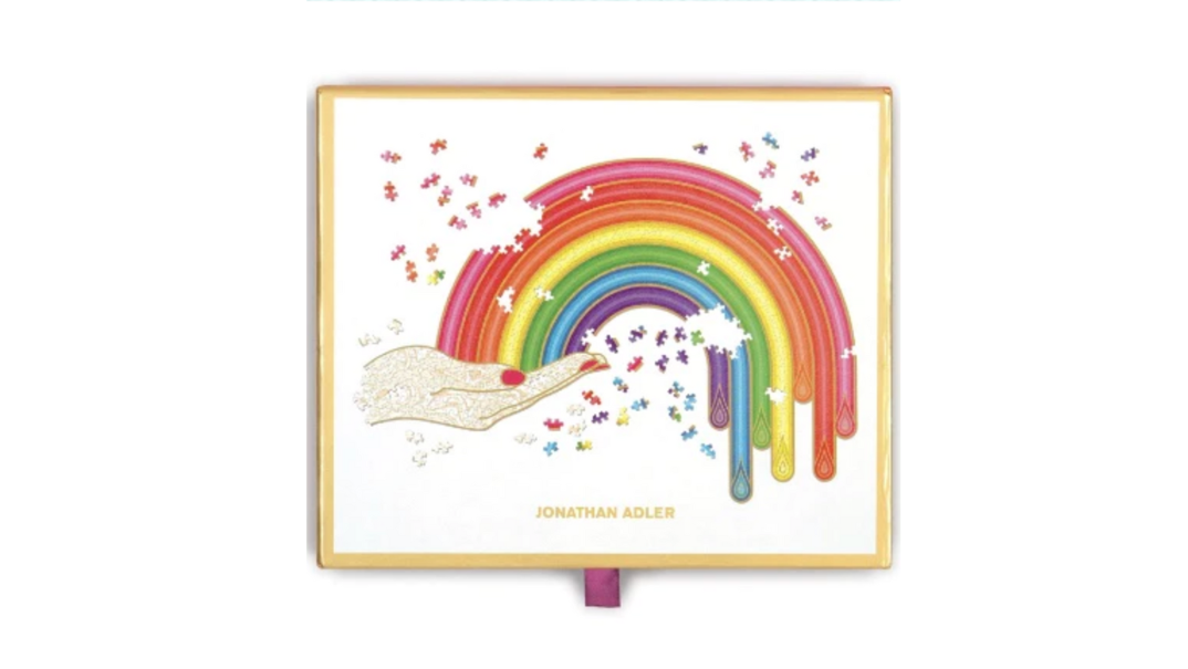 A gold-framed puzzle depicting a red-nailed hand sticking out with a rainbow emerging from its finger tips. The puzzle is mostly complete, with a few pieces out of place laying around it. Gold text reads "Jonathan Adler."