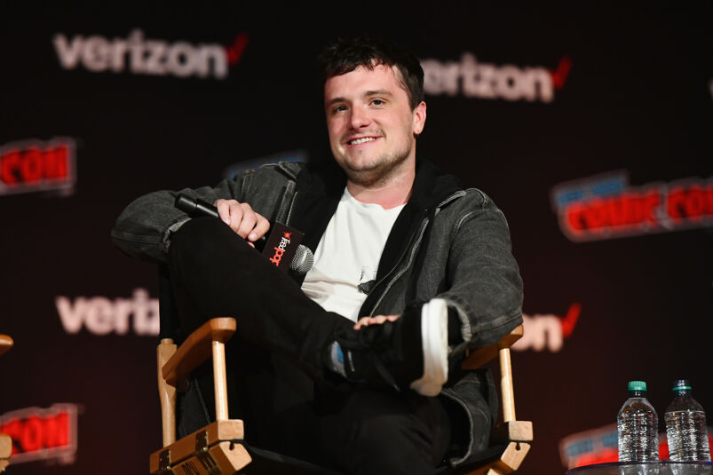 Josh Hutcherson sits in a black director's chair with his legs crossed smiling. He sits in front of a black wall that has Verizon and Comic Con's logo. He wears a gray hoodie over a white t-shirt, black jeans, and black skater shoes while holding a microphone. To his left are two bottles of water.
