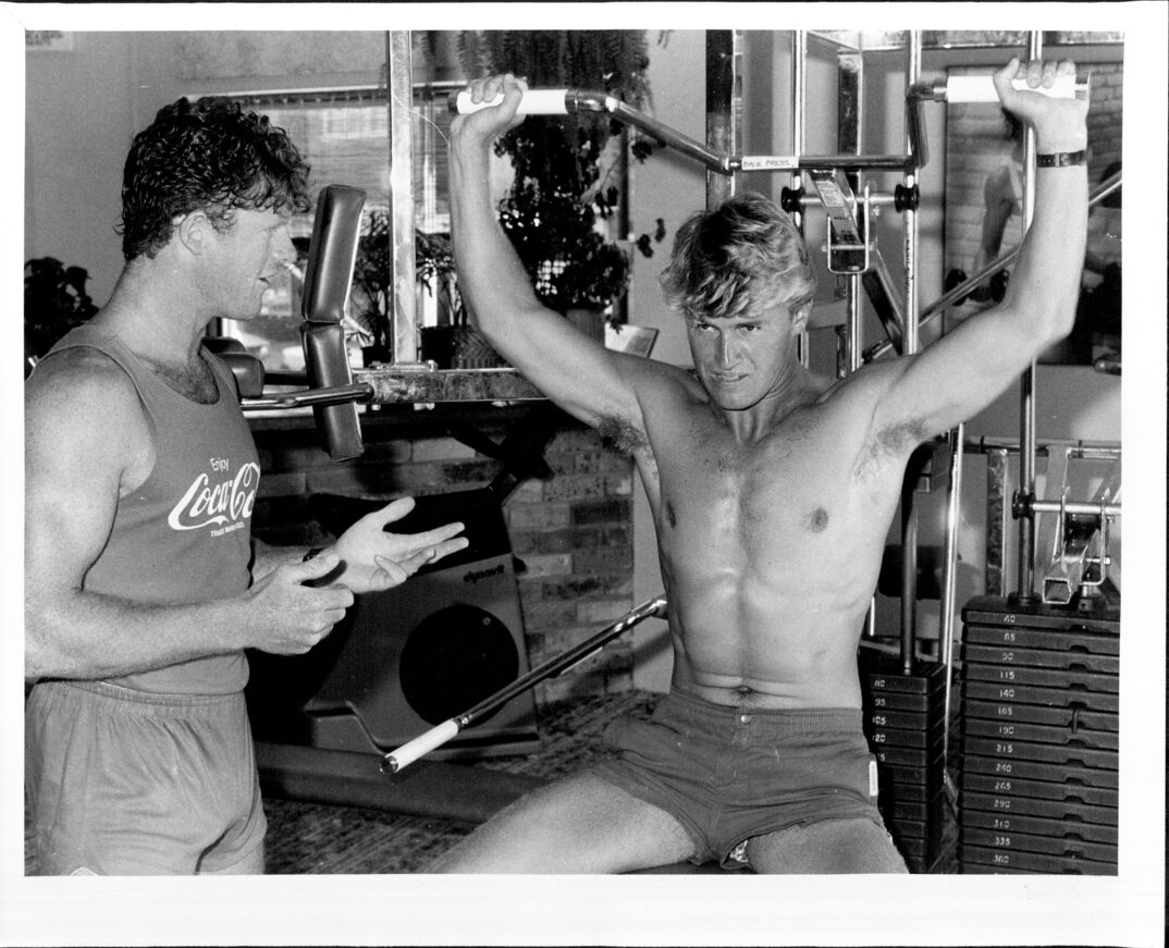 Black-and-white image. A blonde and muscular shirtless man in shorts sits working out his back on a machine in a gym. A dark haired muscular man, in a Coca Cola tank top and tight shorts, stands to his right, talking him through the workout. Behind them, various workout machines sit.