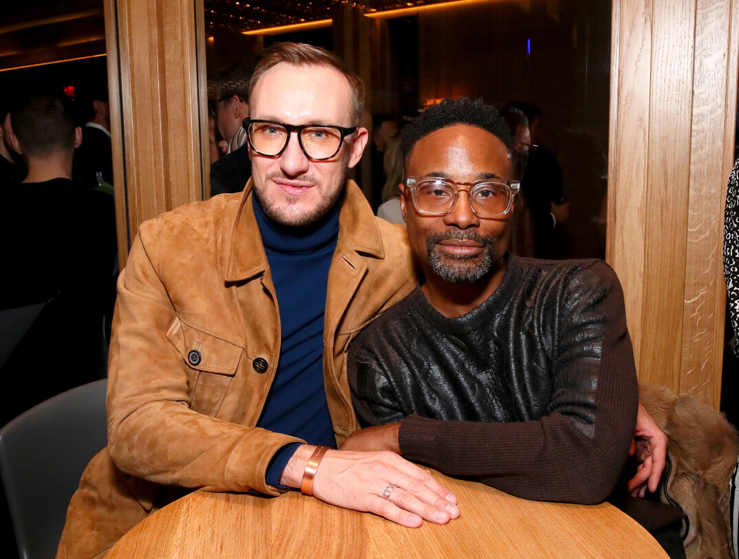 Adam Smith, a white man with thin blonde hair styled in a faux hawk, a thin blonde beard, and thin rimmed black glasses, smiles wearing a navy turtleneck and collared tan leather jacket. He sits at a round wooden table with his arm around Billy Porter, a Black man with curly dark hair, a gray and black beard, and translucent-rimmed glasses. Porter wears a sparkly long sleeve black sweater and leans against Smith, smiling. In the background, nondescript people mill around.