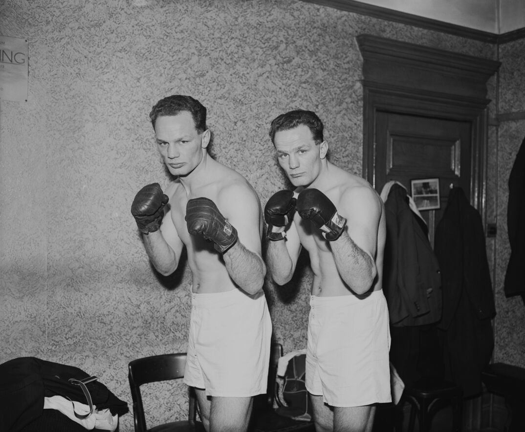 Black-and-white image. Two identical brothers stand in front of a wooden door with coats hanging from it and a few chairs. They are shirtless and stand in white shorts, grimacing as they hold up their fists ready to throw a punch in black wrestling gloves.