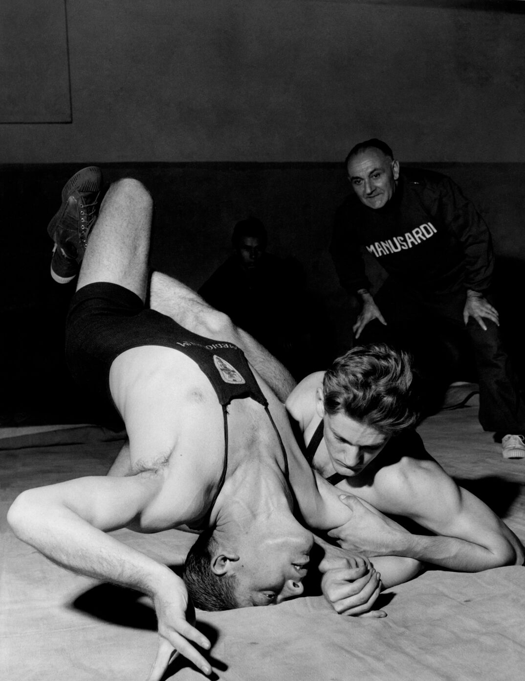 Black-and-white image. On a wrestling mat, two men in tight singlets wrestle with their legs intertwined. Both have blonde hair and muscular builds. Behind them, an older man in a black sweatshirt and sweatpants with a receding hairline watches.