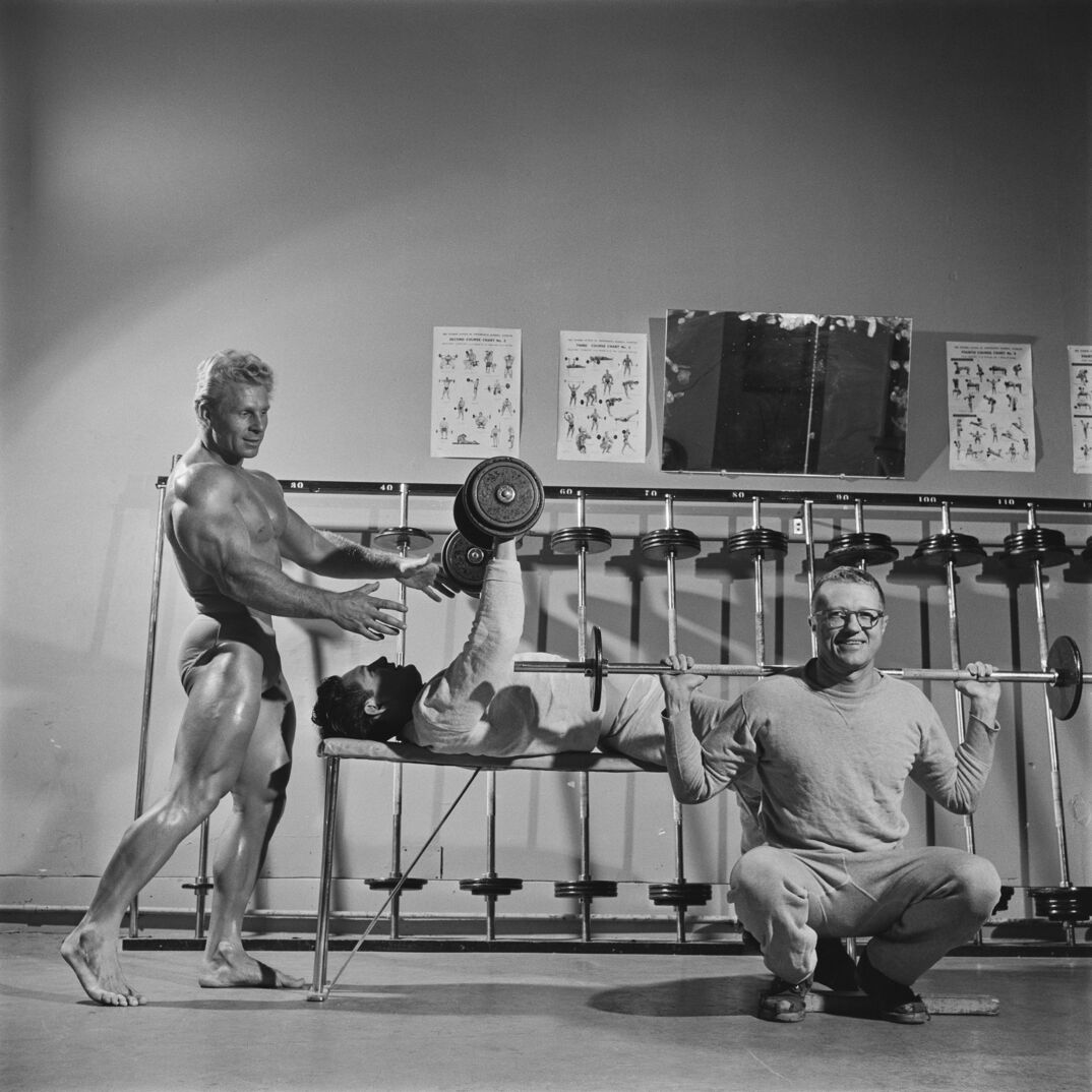 Black-and-white image. A toned and tan blonde man stands in a speedo behind another young man with dark hair who is clothed, laying on his back on a weight bench. The blonde man holds him arms out to spot the dark haired man as he lifts up two weights. In front of them, another young man with glasses, wearing a gray long sleeve and sweat pants, squats and smiles at the camera with a barbel resting on his back. Behind all three men is a shelf with weights and posters modeling workout techniques.
