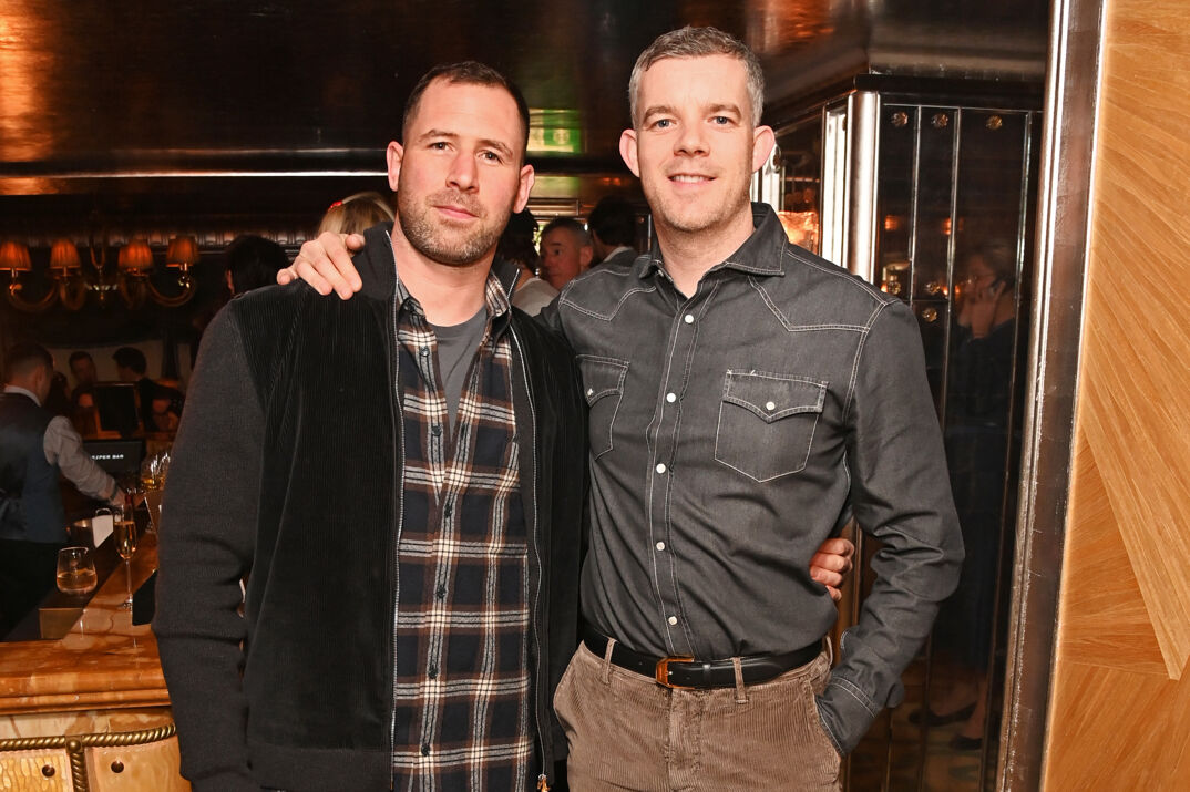 Russel Tovey, a thick built man with short brown hair and a beard, softly smiles wearing a gray tee under a plaid shirt and a black, textured long-sleeved hoodie. He leans against a smiling Steve Brockman, who's slightly taller with salt-and-pepper hair styled in a faux hawk. Brockman wears a collared black shirt tucked into brown corduroy pants and a belt, holding his arm around Tovey. In the background, a nondescript bar with people mulling around.