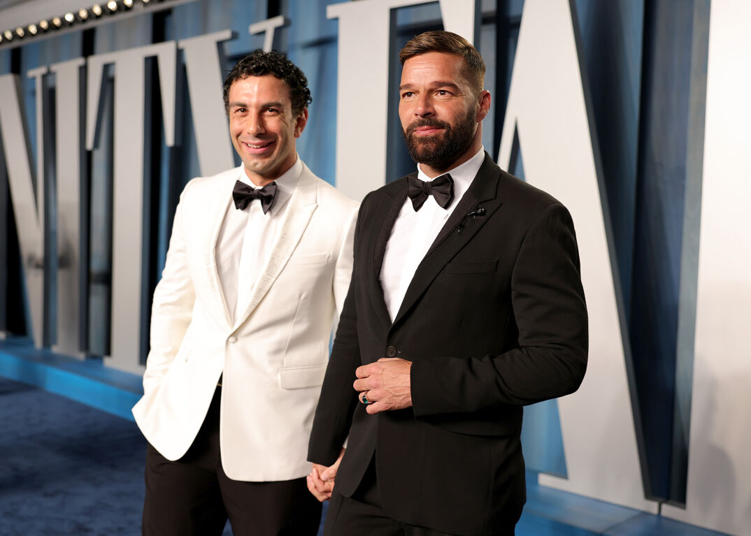 Jwan Yosef, with curly dark hair, stands smiling with his hands in his pockets in a white suit jacket and dress shirt and black dress pants. He has a black bowtie. To his right, Ricky Martin, with dark hair featuring bleached tips and a full brown beard and mustache, stands in a black suit jacket and dress pants. With a ring on his left hand, he adjusts the jacket over a white dress shirt and also wears a black bowtie. The two are on a blue carpet, posing in front of a sign that reads "Vanity Fair."