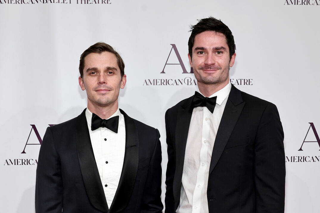 Antoni Porowski softly smiles, wearing his brown hair brushed back and sporting a stubbly mustache and goatee. He stands in a white dress shirt and black suit jacket with a black bowtie. To his right, Kevin Harrington (slightly taller) stands smiling in a matching suit. Harrington has darker hair, tousled on top of his head, and a stubbly beard and mustache. The two are posed at in front of a white step-and-repeat reading "American Ballet Theatre."