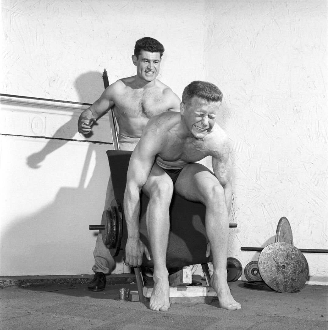Black-and-white image. A muscular shirtless man with dark hair stands behind a weight rack widely grinning. In front of him, a blonde muscular and shirtless man squats in concentration as he works out his bulging biceps. He wears a black speedo and no shoes, leaning against the bench loaded with weights. A few spare weights and barbels rest against the wall.