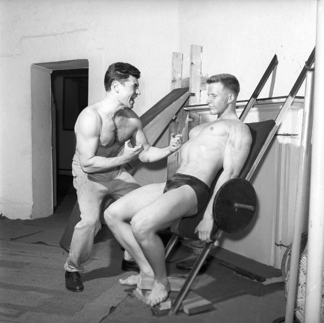 Black-and-white image. A muscular shirtless man with a hairy chest stands to the right of a blonde muscular shirtless man in a black speedo. His back is against a weight bench as he cheerfully works out his legs by leaning against it. He is barefoot.
