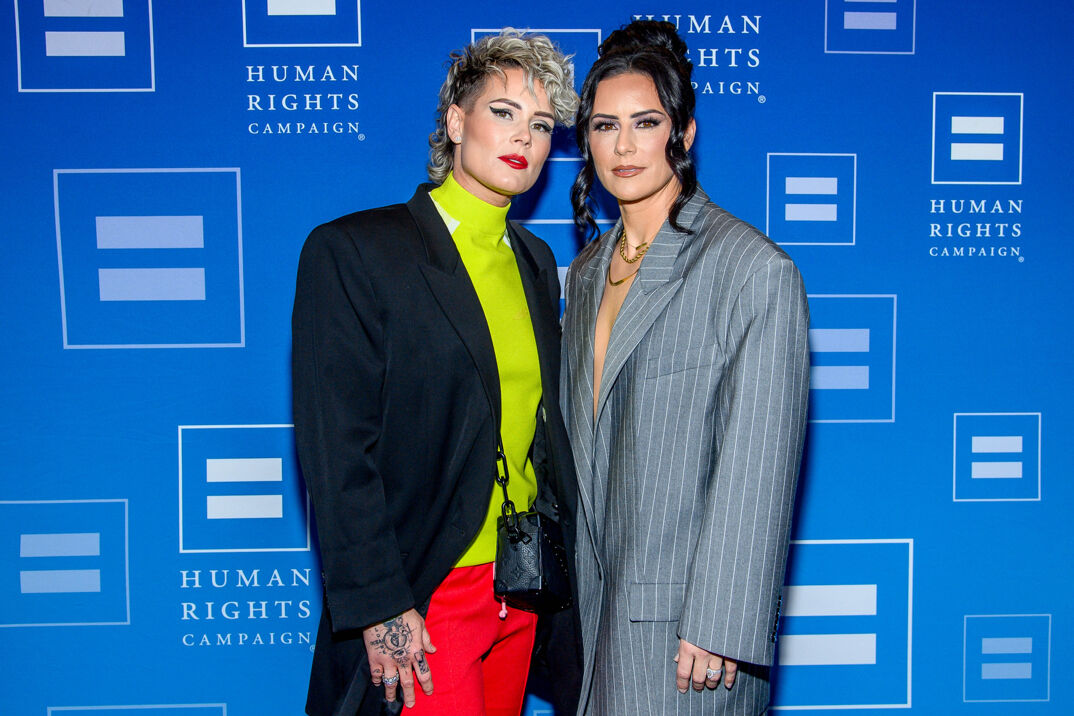 Ashlyn Harris, with dark red lipstick, thick brows, and cat-eye curved eyeshadow, looks into the camera mysteriously. She sports a bleached blonde mullet, wearing a neon yellow turtleneck under a purple suit jacket and red slacks. Her right hand hangs out of the sleeve, revealing tattoos over her fingers. She stands next to Ali Krieger, with long brown hair split into locks on either side of her face and tied behind her head in a bun. Krieger wears a loose gray pinstriped suit jacket. The women are posed in front of a blue step-and-repeat reading "Human Rights Campaign."