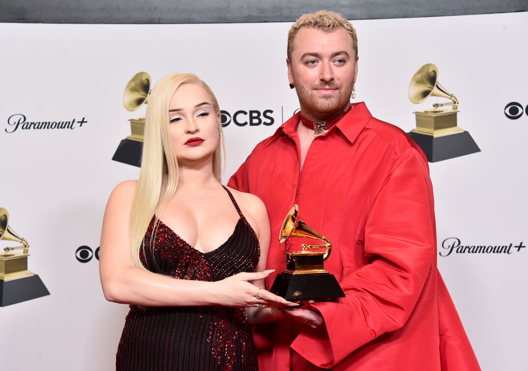 Kim Petras, with dark red lipstick, long bleached blonde hair and wearing a busty maroon dress, smiles alongside Sam Smith (in an oversized and collared long sleeved red dress). They both hold a golden gramophone Grammy Award in front of a step-and-repeat.