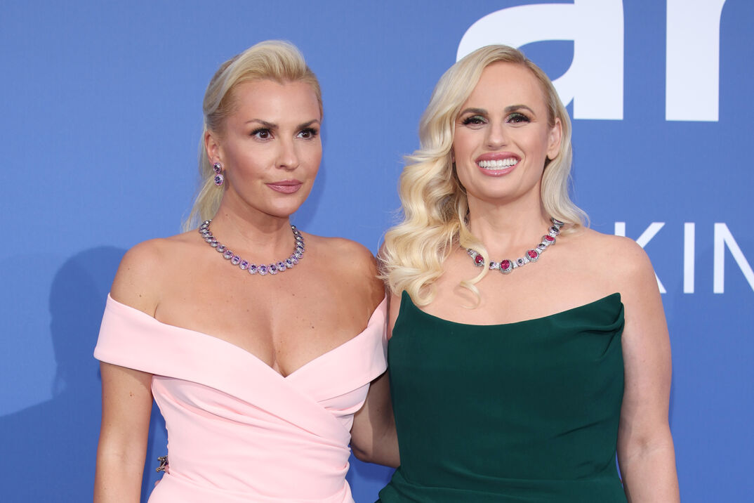 Ramona Agruma, with long blonde hair tied behind her head and pink lipstick, wears a low cut and sleeveless light pink dress revealing her cleavage and a purple necklace. She stands with her arm around fiancee Rebel Wilson, same height, who has curly blonde hair hanging over her shoulders. Wilson has a huge smile and wears a sleeveless black dress. The two are posed on a red carpet in front of a blue step-and-repeat.