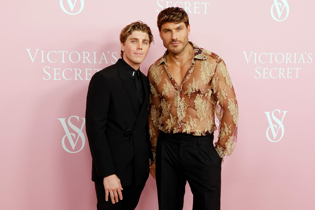Lukas Gage, with wet-looking dirty blonde hair, smiles softly at the camera in an all black suit, resting his hand on his right leg. Next to him stands Chris Appleton, who's slightly taller with textured long dirty blonde hair. Appleton wears a sheer floral dress shirt, revealing a muscular body underneath, tucked into tight-fitting black pants. The men are posed in front of a pink step-and-repeat reading "Victoria's Secret."