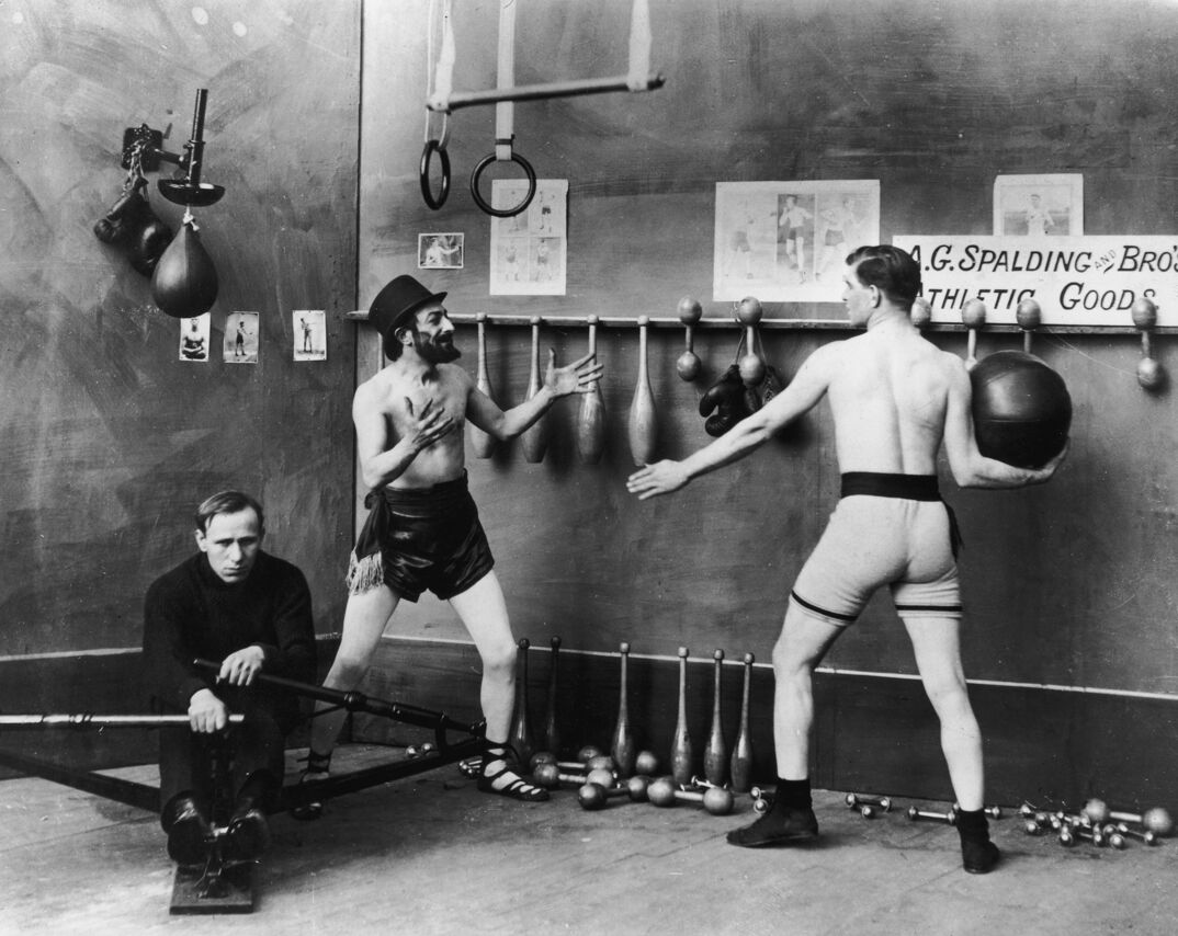 A shirtless bearded man in a top hat and loose black shorts holds out his hands. Across from him, another shirtless man in tight gray shorts winds up to throw a black medicine ball at him. They stand in front of a wall filled with weights and rings, featuring a sign that says "A.G. Splading and Bros Athletic Goods." To their right, a fully clothed young man in a long sleeve black shirt and pants sits on a rowing machine.