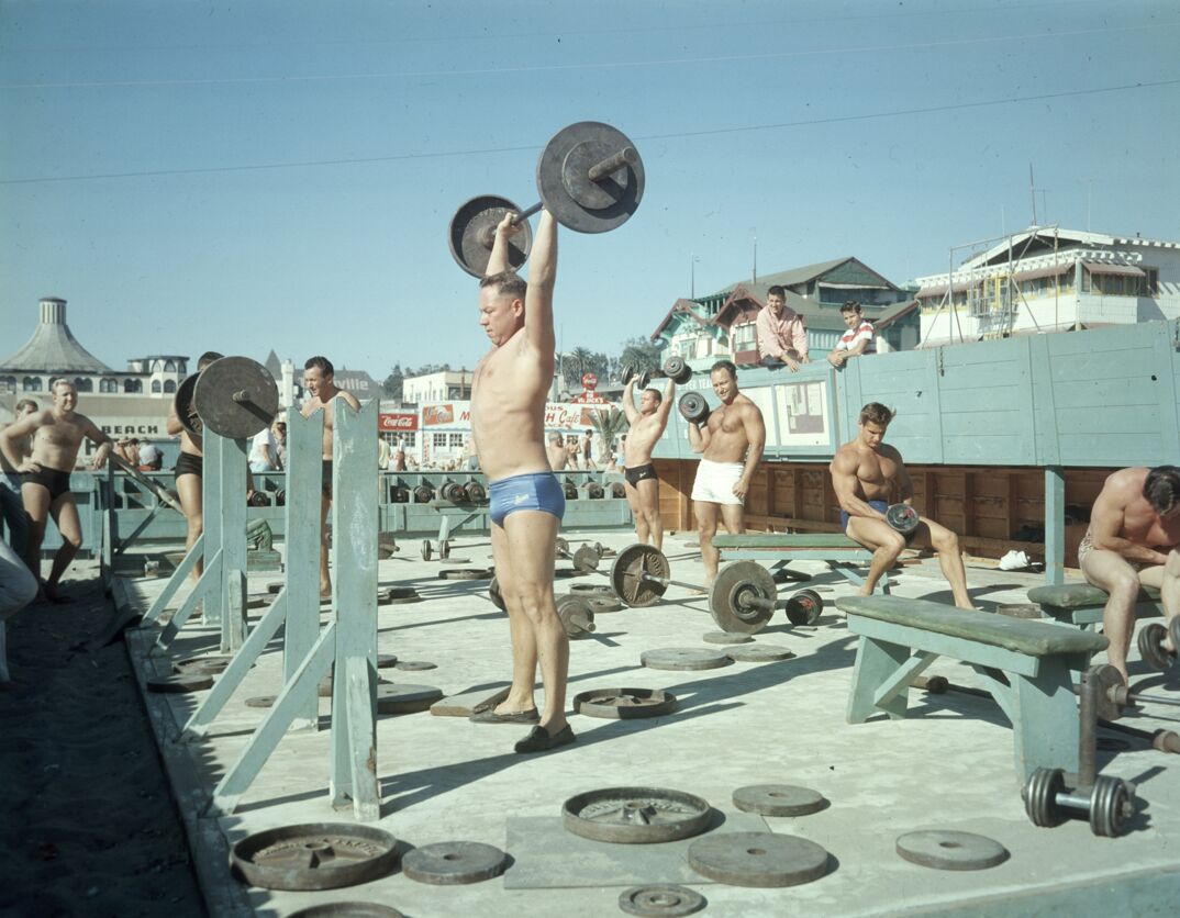 On a concrete floor, surrounded by little wooden beach huts featuring signs like "BEACH" and "Coca-Cola," a shirtless man stands in the center of the image wearing a blue speedo and loafers. He holds a heavy barbell above his head in front of a wooden weight rack. Around him, other shirtless men sit around in speedos and shorts, working out their biceps with weights, flexing and watching in admiration. Weights sit strewn around the wooden benches and racks.