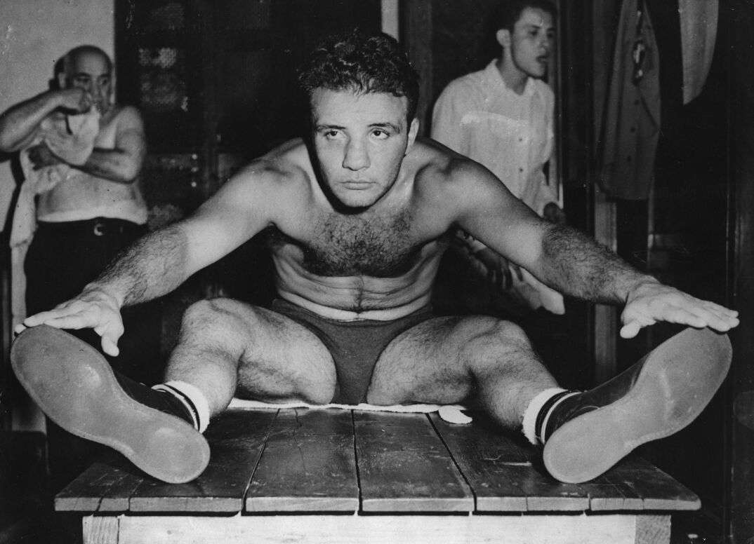 Black-and-white image. A shirtless man with a hairy chest leans forward and touches his toes. He wears gray briefs, white socks, and dark shoes. He looks off from the camera, sitting on a wooden table. In the background, an older man puts on a tank top while sitting down and a younger man in a white dress shirt walks by.