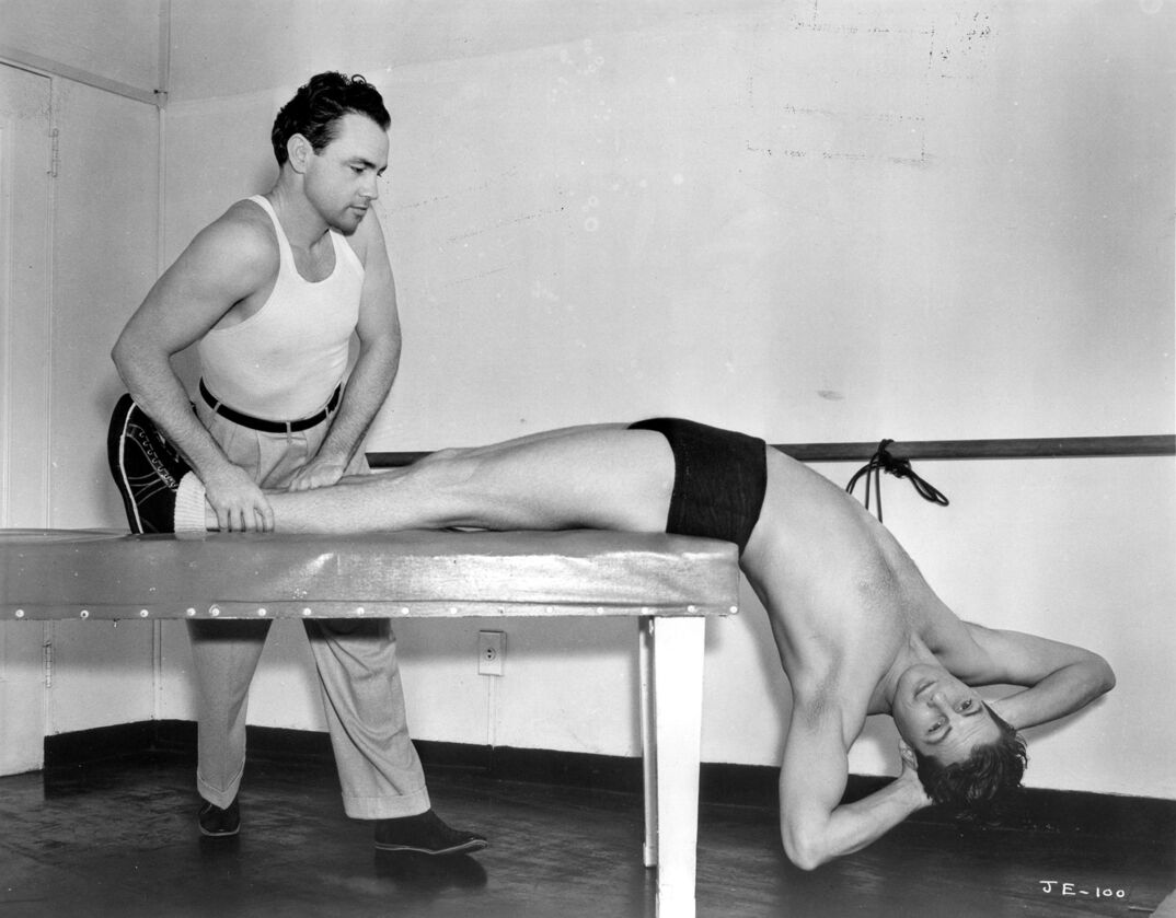 Black-and-white image. On a cushioned bench, a tall and smooth young man rests his muscular legs on the bench and hangs his torso off. He is shirtless in black briefs, and holds his hands behind his head. On the left of the bench, a muscular man with dark hair in a white tank top holds down his legs for the workout. They stand in a white room with a bar around the edges.