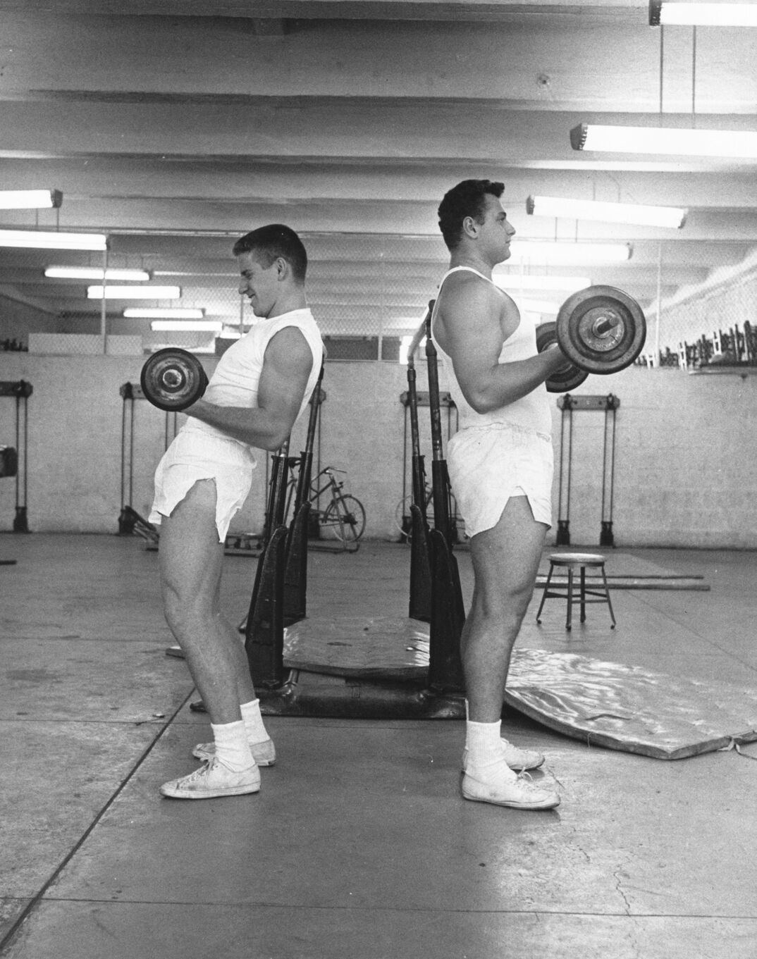 Black-and-white image. In a gym with brick walls and overhead lighting, two men in white tank tops and shorts stand back to back holding barbells in their hands to bicep curl. The taller man on the right stands straight while the shorter man on the left hunches his back. Behind them are mats, weight racks, and a bicycle.