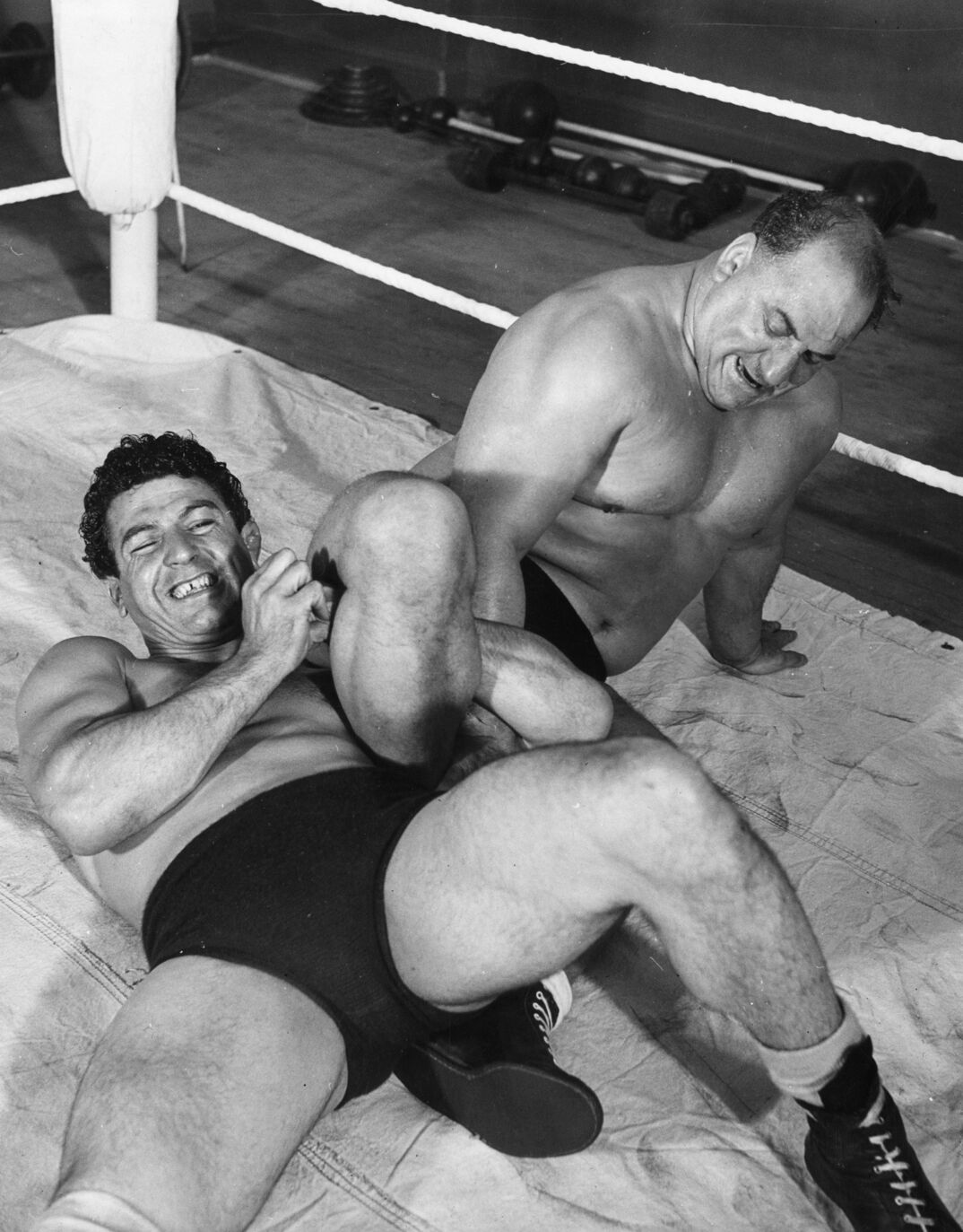 Black-and-white image. In a white wrestling ring, a young shirtless man with curly hair, wearing tight black shorts, lays on his back. His left bicep is wrapped around the leg of another muscular shirtless man on his left as the two wrestle.