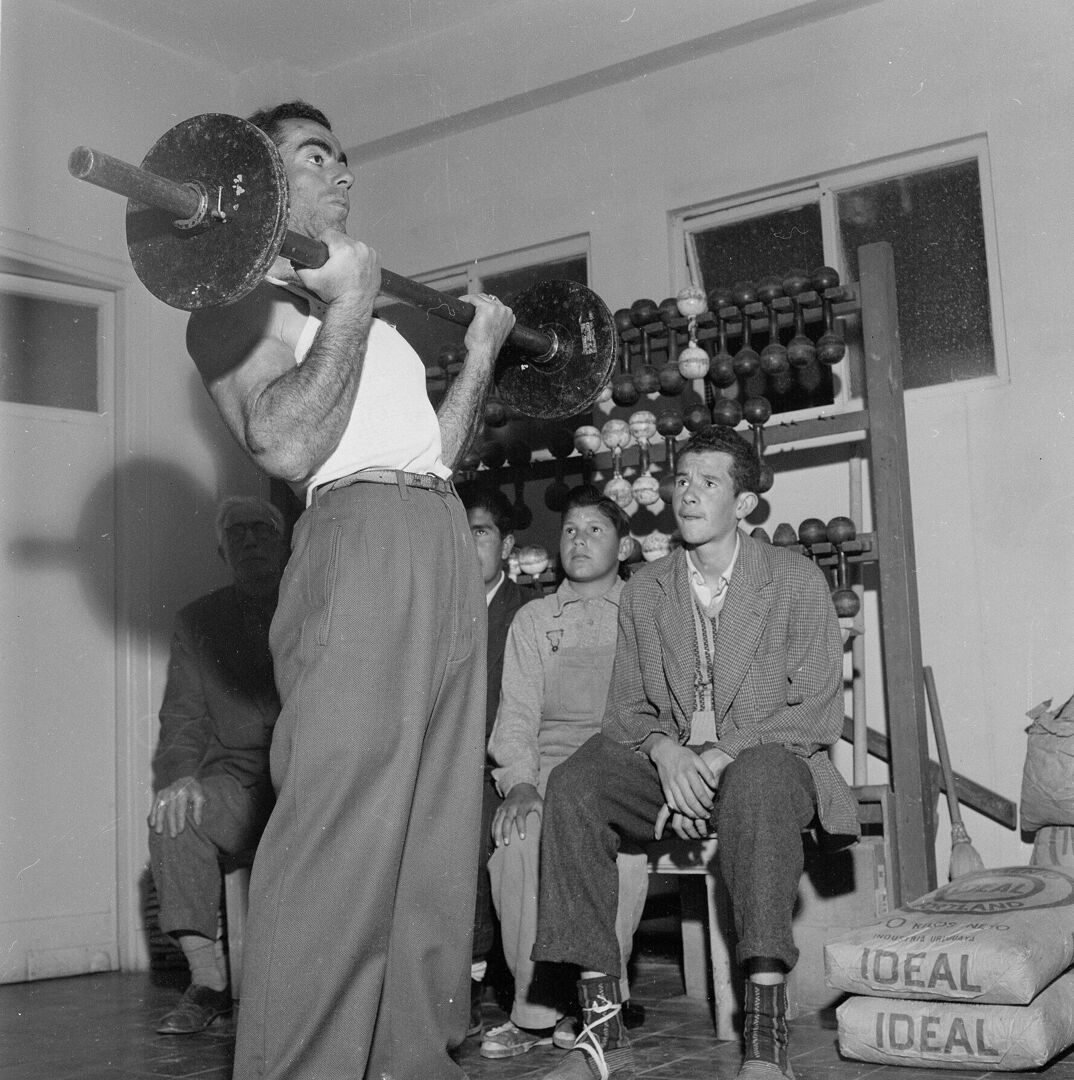 Black-and-white image. A tall man in loose pants and a white tank top stands with a barbel in his arms, modeling a bicep curl. Behind him, young men watch his form from a bench. They sit in front of two windows and a shelf filled with barbels and weights. 
