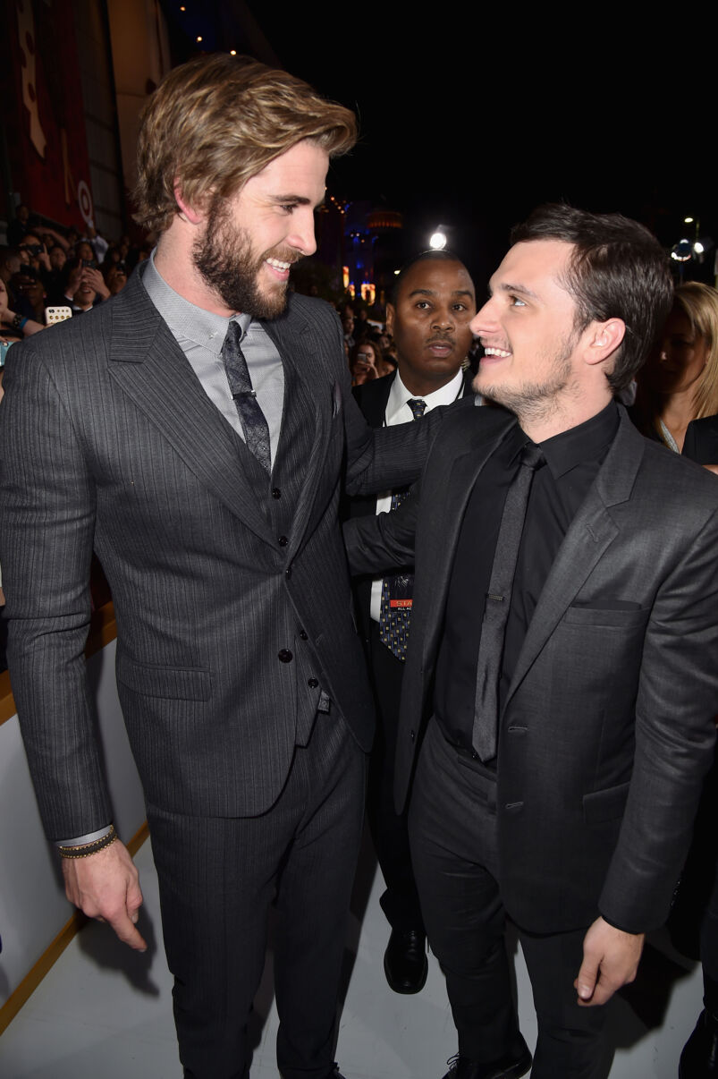 Josh Hutcherson, wearing a black suit jacket over a black dress shirt tucked into tight-fitting black slacks and a black tie, stands with his arm around Liam Hemsworth, who is a head taller than him. Hemsworth, with blonde hair and a full blonde beard, wears a slim gray striped suit over a light gray shirt and paisley gray tie. They look intently into each other's eyes smiling on a red carpet, with a crowd of nondescript fans and security guards standing behind them.
