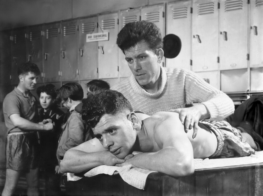 Black-and-white image. In a locker room, a man in a white sweater looks off as he massages the shirtless back of another young man with dark hair. The young man lays on his stomach on top of a wooden table covered by a towel. He thoughtfully rests his chin on top of his hands. 