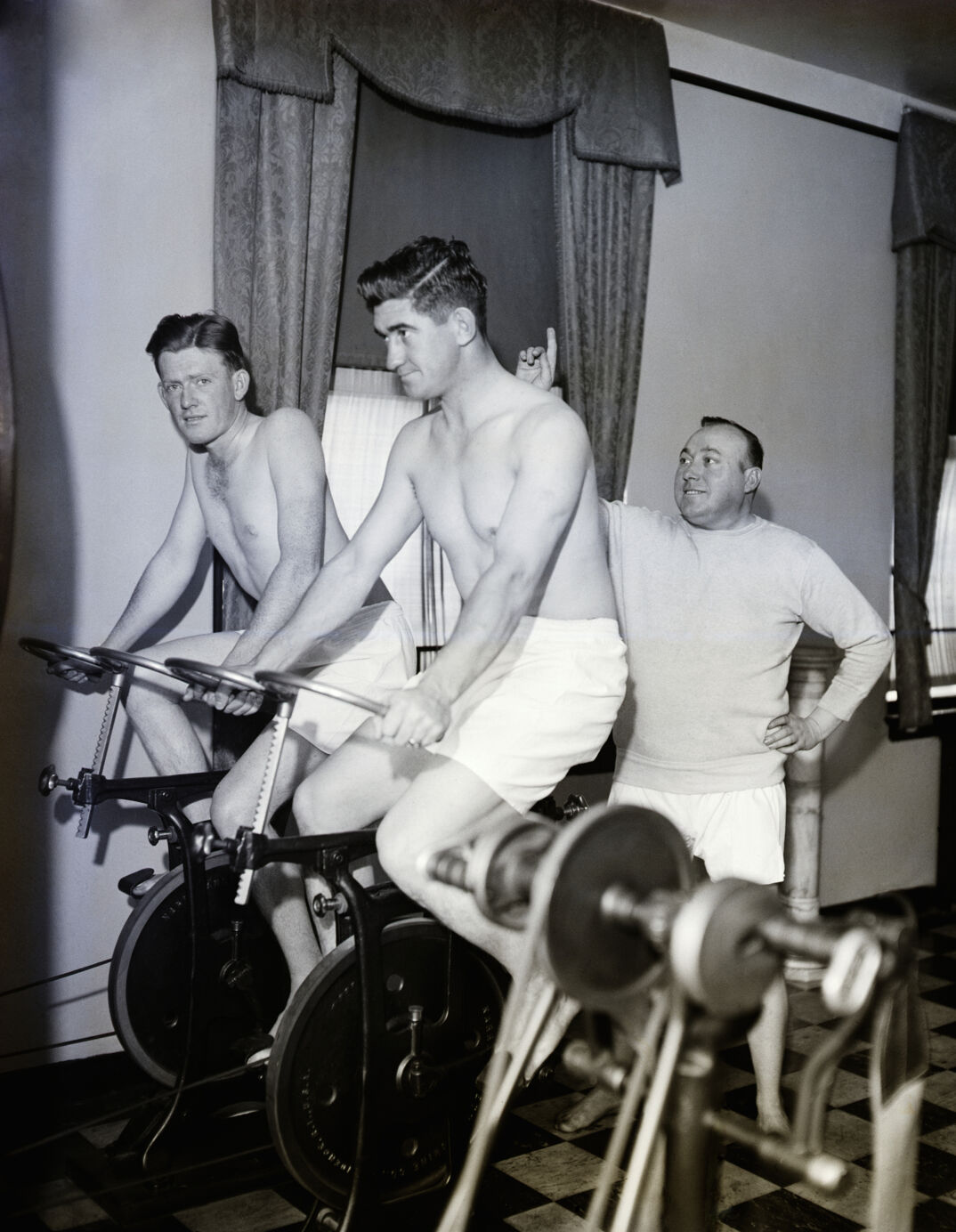 Black-and-white image. Two shirtless and fit young men are on workout bikes and sit up straight as they pedal. They are in a room with two curtained windows and other workout machines. Behind them, a shorter and stout man in a white long sleeve shirt and shorts points and leads them in their workout.