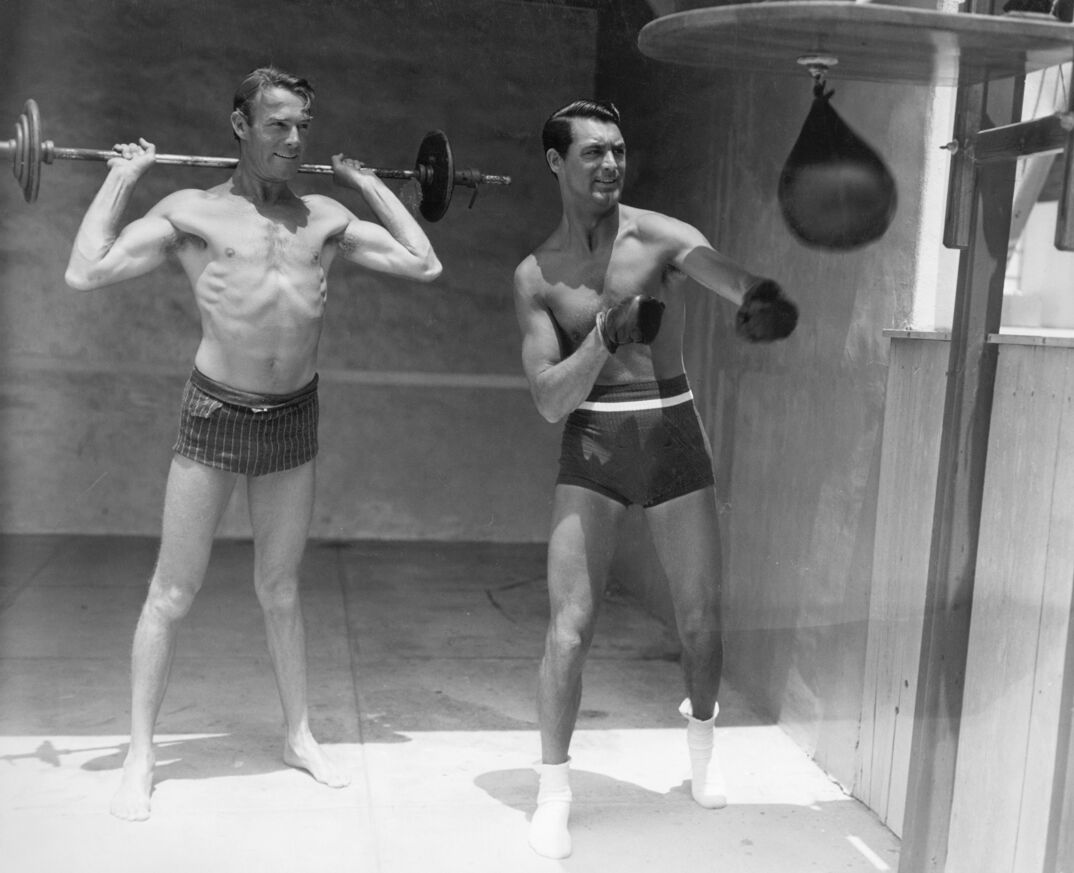 Actors Randolph Scott (L), and Cary Grant show off their athletic prowess. Scott lifts weights while Grant punches at a speedbag.