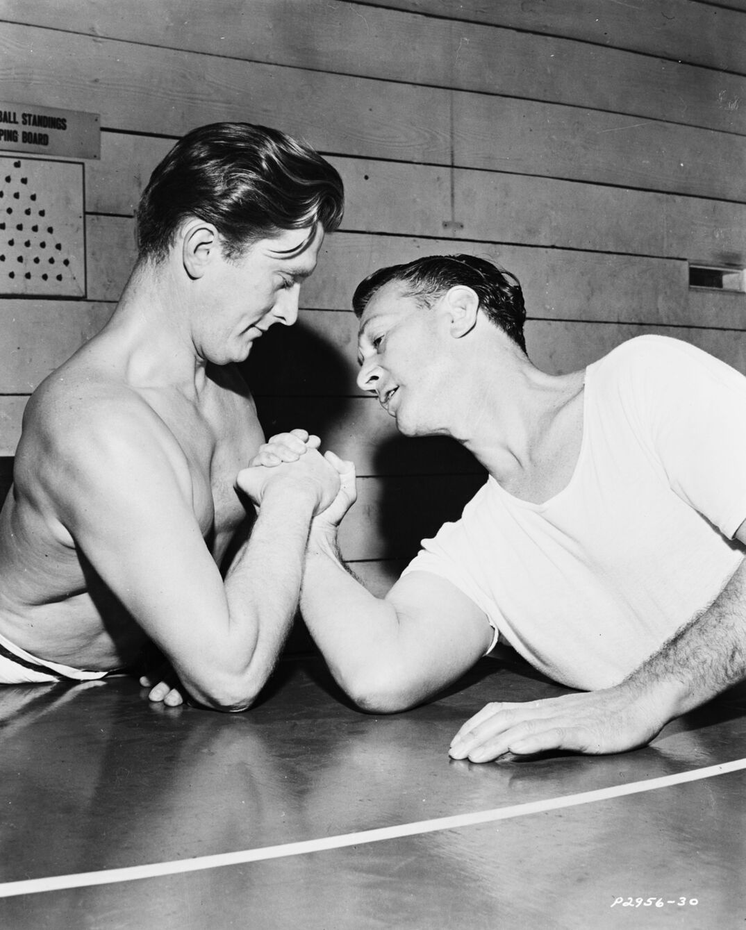 Black-and-white image. In a wood paneled room, two men rest their arms on a sleek table armwrestling. The man on the left is shirtless with slicked back hair. The man on the right wears a white t-shirt and rests his other arm on the tabletop for leverage.