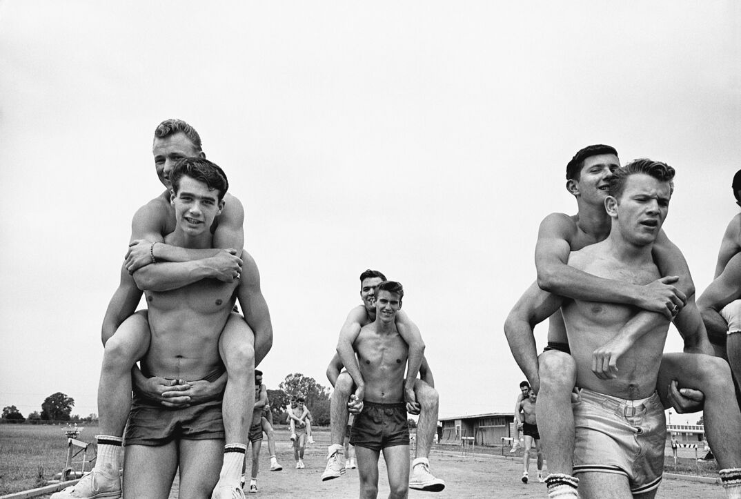 Black-and-white image. On a dirt track with trees in the background, groups of shirtless men walk around in shorts giving other shirtless young men piggy back rides. There are 6 different couples of men throughout the frame walking around.