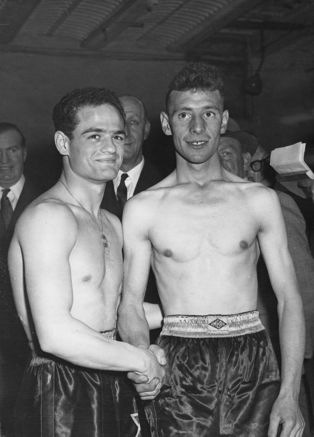 Black-and-white image. Two shirtless wrestlers stand wearing high-waisted gym shorts and shaking hands, smiling for the camera. The man on the left is shorter with dark hair. The taller man to the right has blonde hair and smiles with his teeth. Behind them, an assortment of men in suits watch in interest.