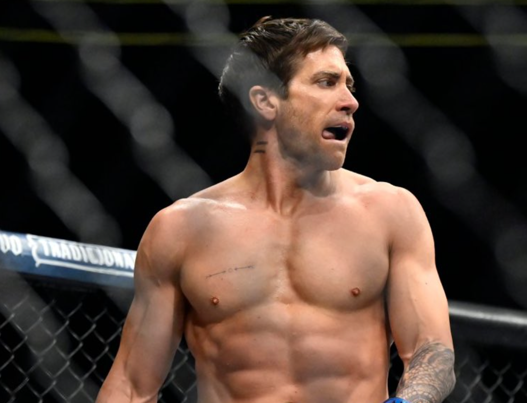 Jake Gyllennhaal stands shirtless and licking his lips in a wrestling ring in a still from upcoming movie 'Road House.' He is muscular with tattooed forearms and an indiscernible text tattoo on his right pec.