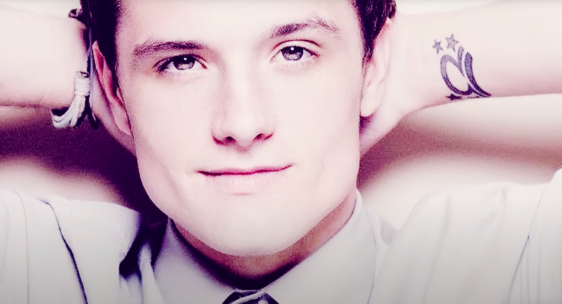 Josh Hutcherson smiles softly with his hands behind his head. He is wearing a white button-down shirt that continues off frame. His hair is out of frame, however a star sign tattoo and a bracelet are visible on his wrists.