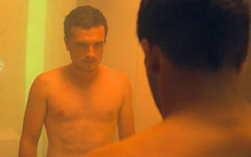 In a scene from 'Future Man,' Josh Hutcherson stands wet in a yellow-hued room naked. He has chest hair and a left nipple piercing. His character looks menacingly at his clone, pictured from the behind, in a stare-off.