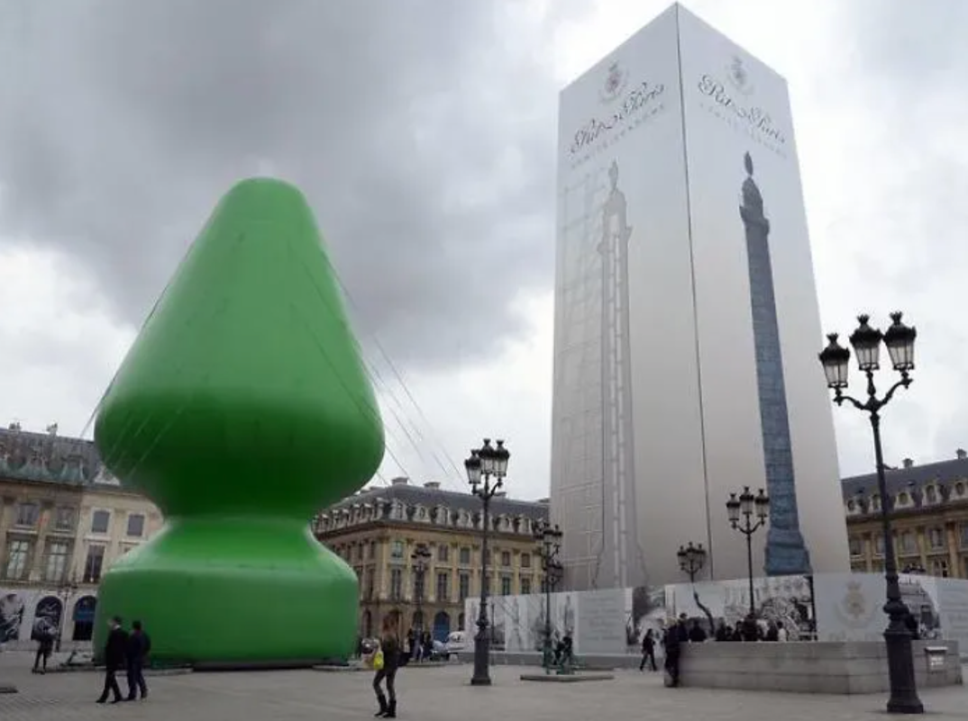 In an outdoor square, with passerby walking through and tall, black street lamps, rests a giant green minimalistic Christmas tree that resembles a butt plug.
