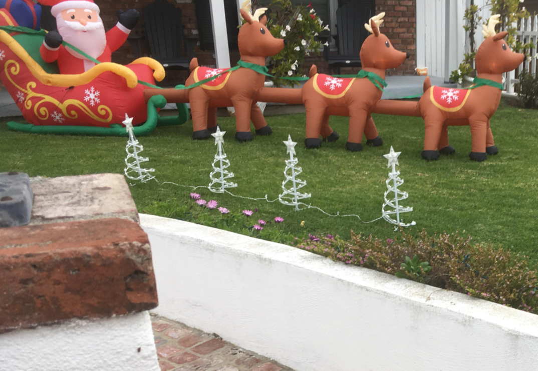 A grassy front yard decorated for Christmas at daytime. An inflatable Santa, in a red coat and hat, sits waving in a sled. He's led by three brown reindeer with snowflake-adorned saddles. The four legged reindeer are posed one in front of the other, connected by what appeared to be a brown leg inserting into the rear of the reindeer in front of them.