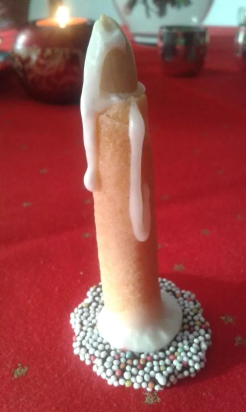 A tan colored candle sits glued on top of a red, green, and white buttoned pad on a red tablecloth. The tan colored candle is dripping in what appears to be white hot glue, resembling a phallus.