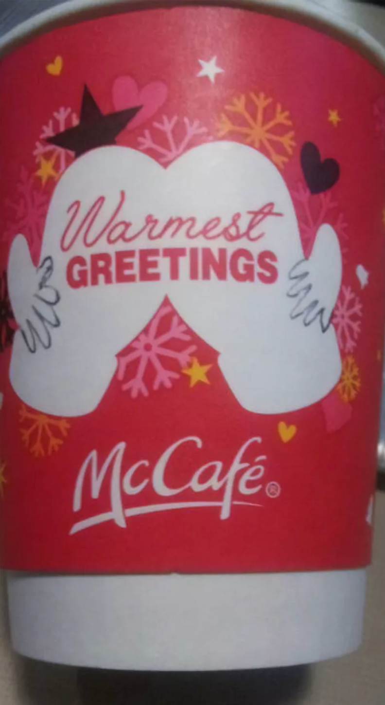A white coffee cup with a heat sleeve from McCafe. The red sleeve reads "Warmest Greetings" on two white mittens being held by hands, resembling a butt being spread open. Around the unfortunate illustration are hearts, stars, and snowflakes.