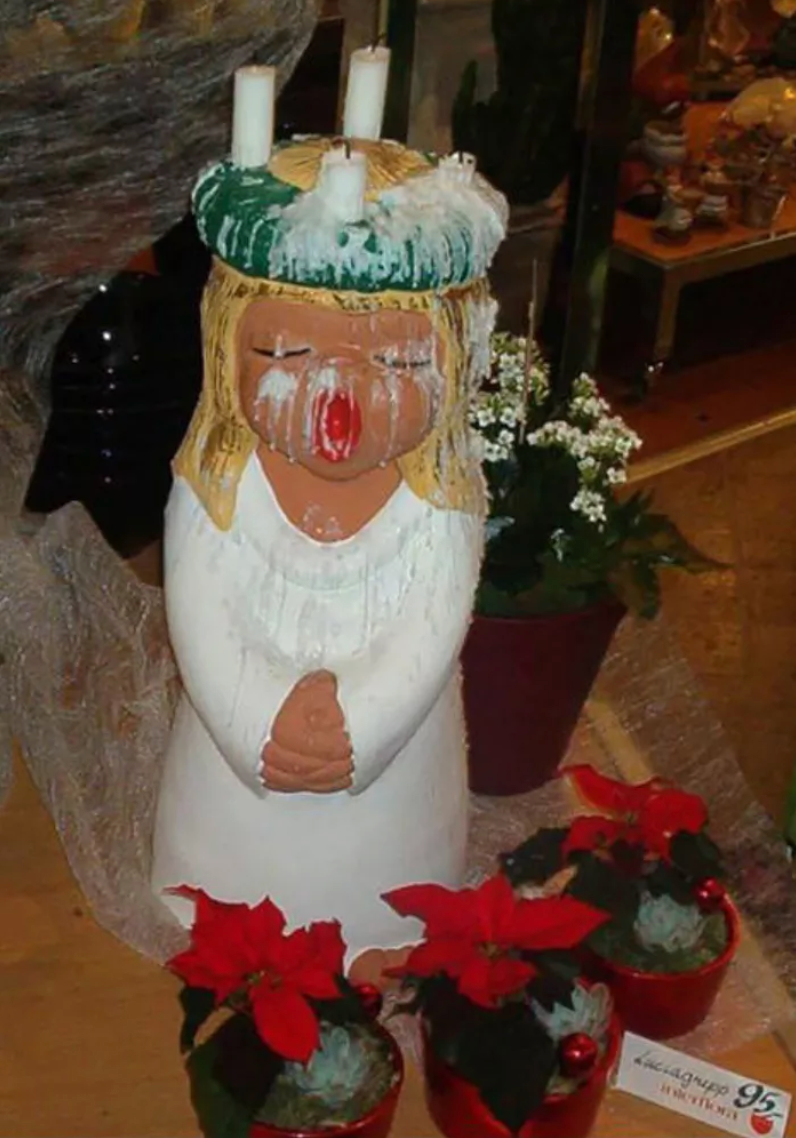 A ceramic decoration of a blonde angel wearing a white dress singing with her mouth open. On her head, four white candles have melted, leaving strips of white dripping down her face. She's surrounded by poinsettia plants in a nondescript living room tableau. 