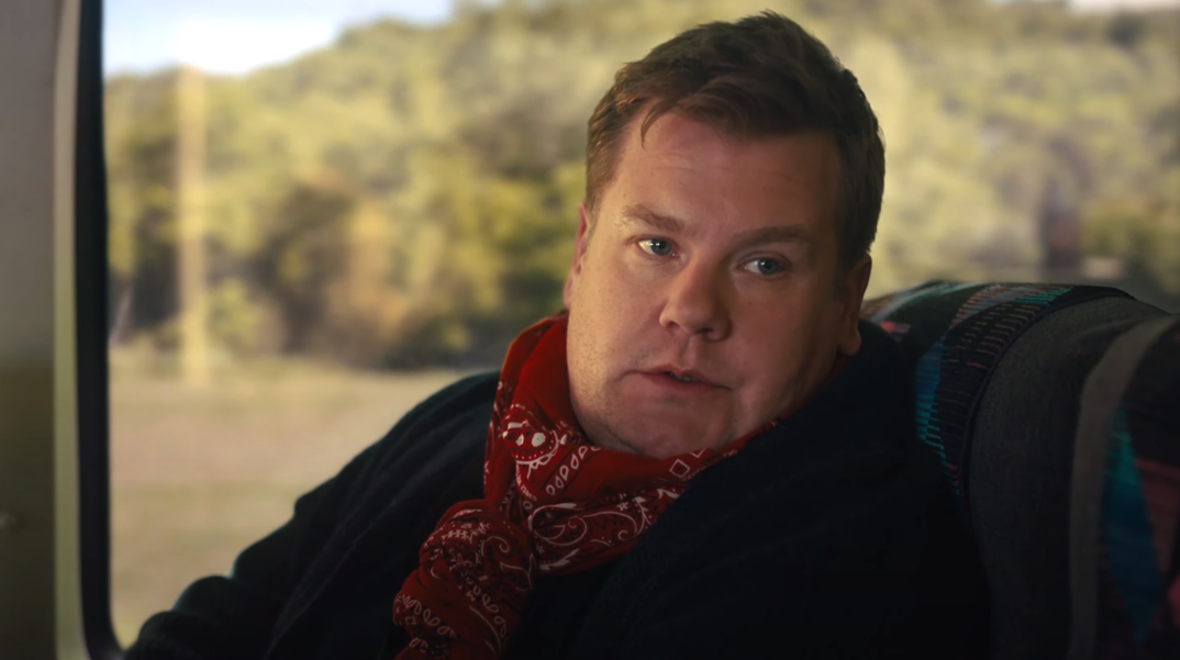 James Corden, wearing a red bandana around his neck and a dark blue sweater, sits in a blue bus seat as green woods are pictured outside the window. The image is a still from the movie 'The Prom.'