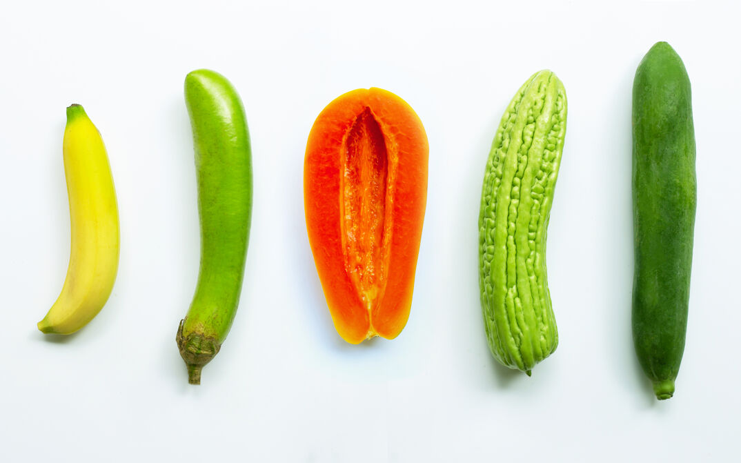 A banana, long green eggplant, ripe papaya cut open, bitter melon, and green papaya lay on a white tabletop and are pictured from a bird's eye view.